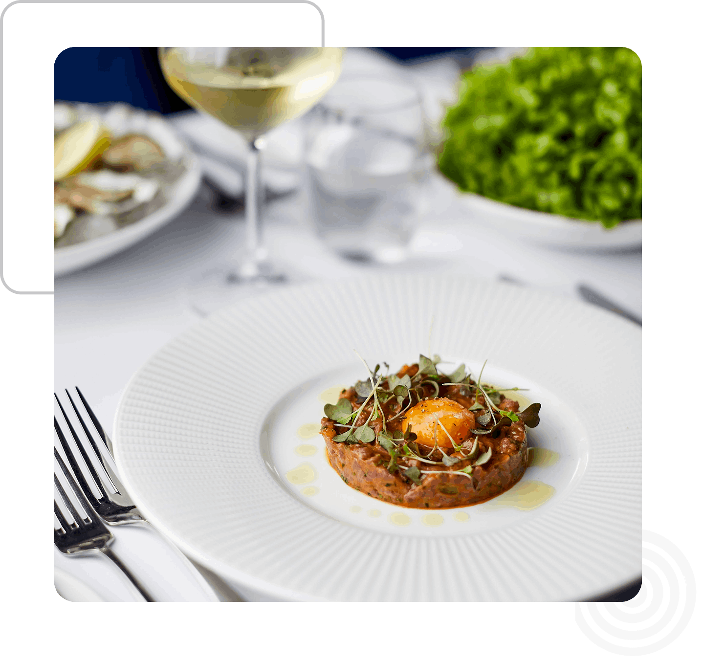 A plated dish with a central mound of tartare topped with a small egg yolk, garnished with microgreens, on a white plate. A fork and knife are on either side, with a glass of white wine nearby.