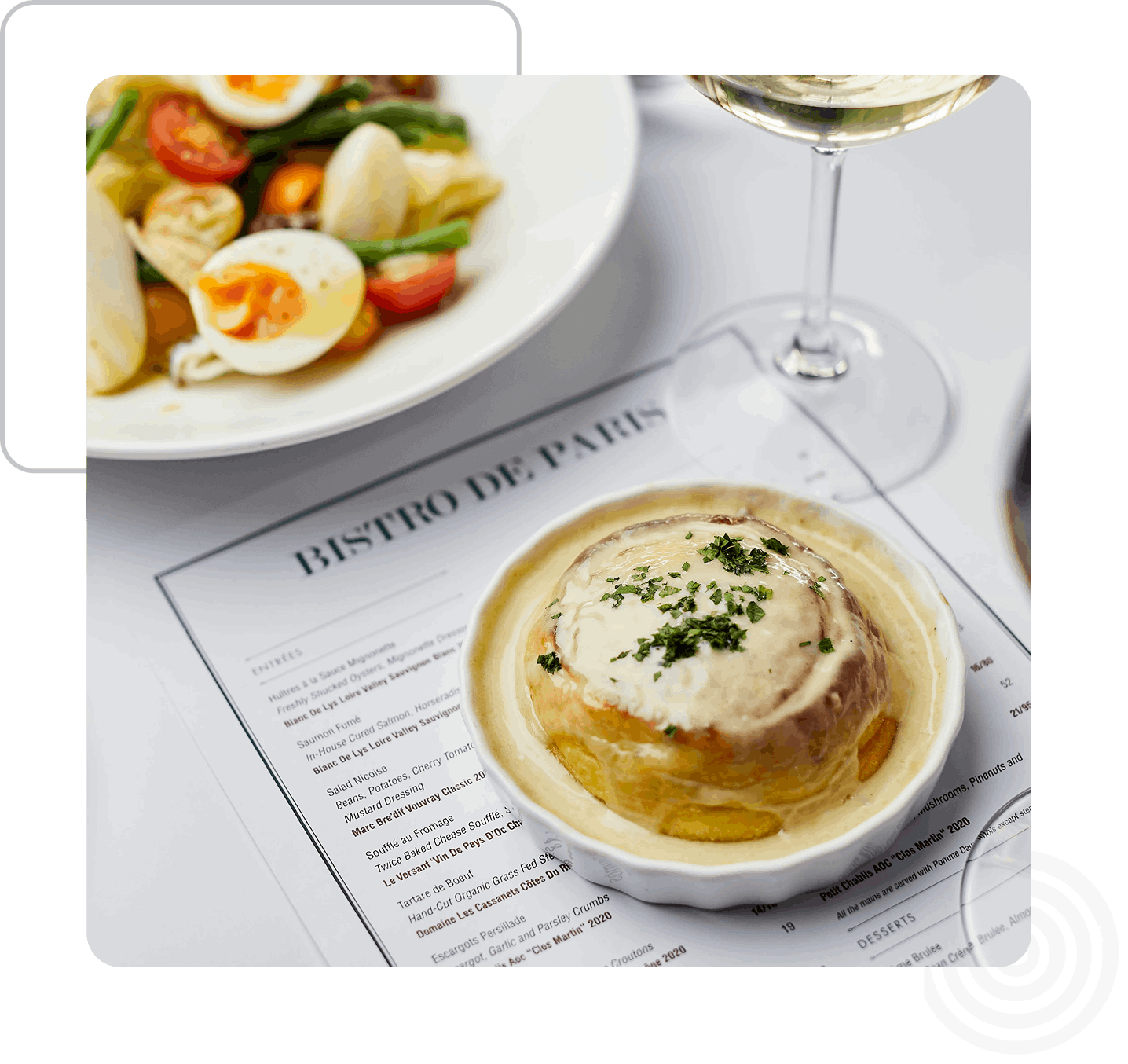 A table setting with a savory pastry in a dish on a menu, a salad with eggs and vegetables, and a glass of white wine.